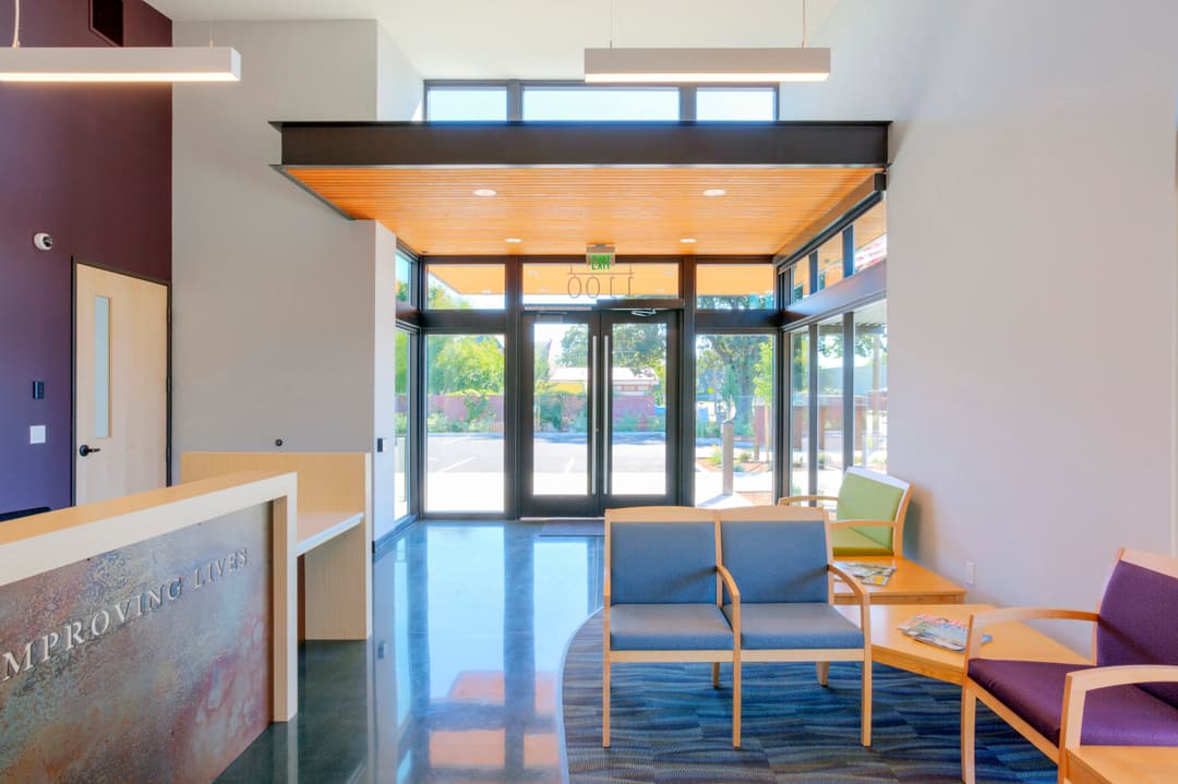 Modern reception area with wooden accents, seating, and large glass entrance. Bright and welcoming.