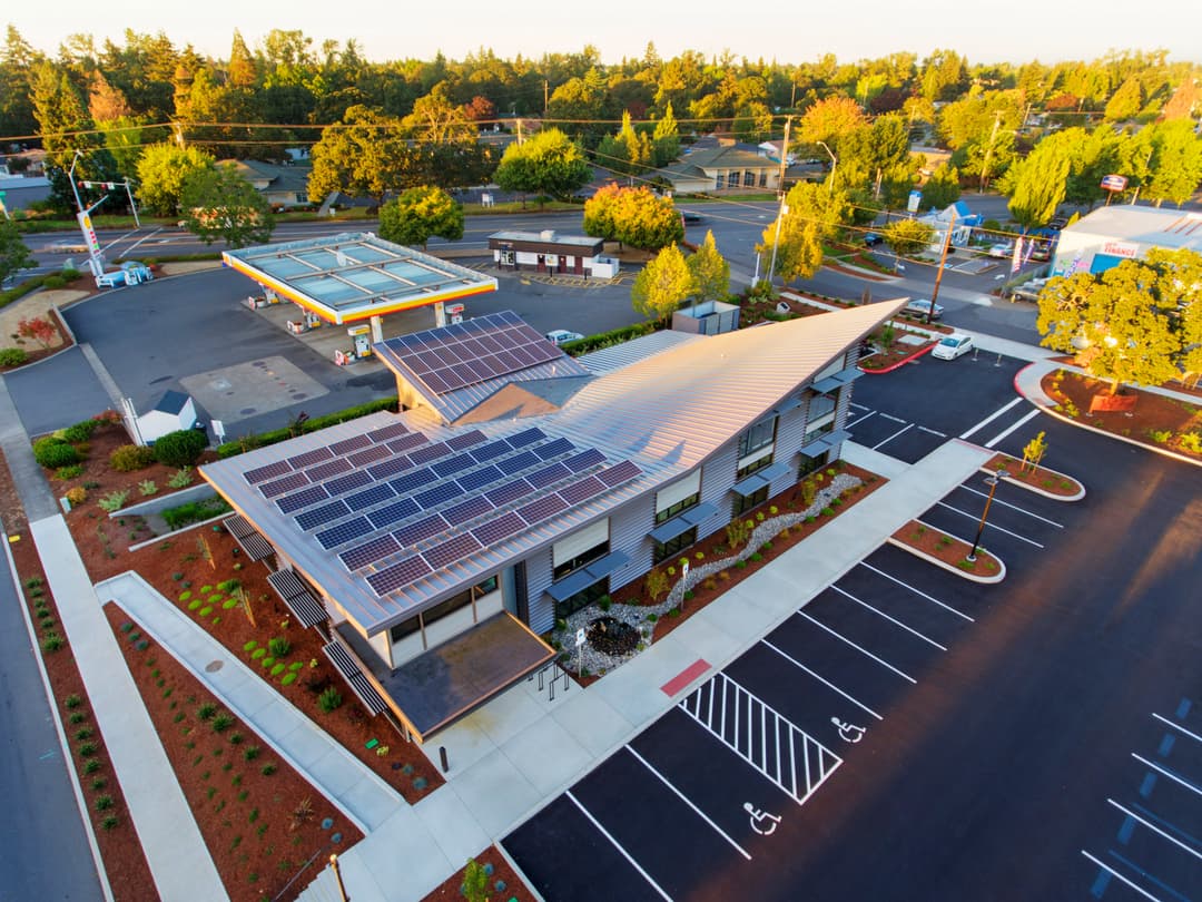 Aerial view of a modern building with solar panels, surrounded by greenery and parking spaces.