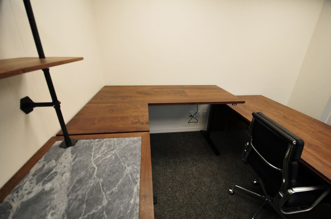 Modern office workspace featuring wooden L-shaped desk and black chair on carpeted floor.
