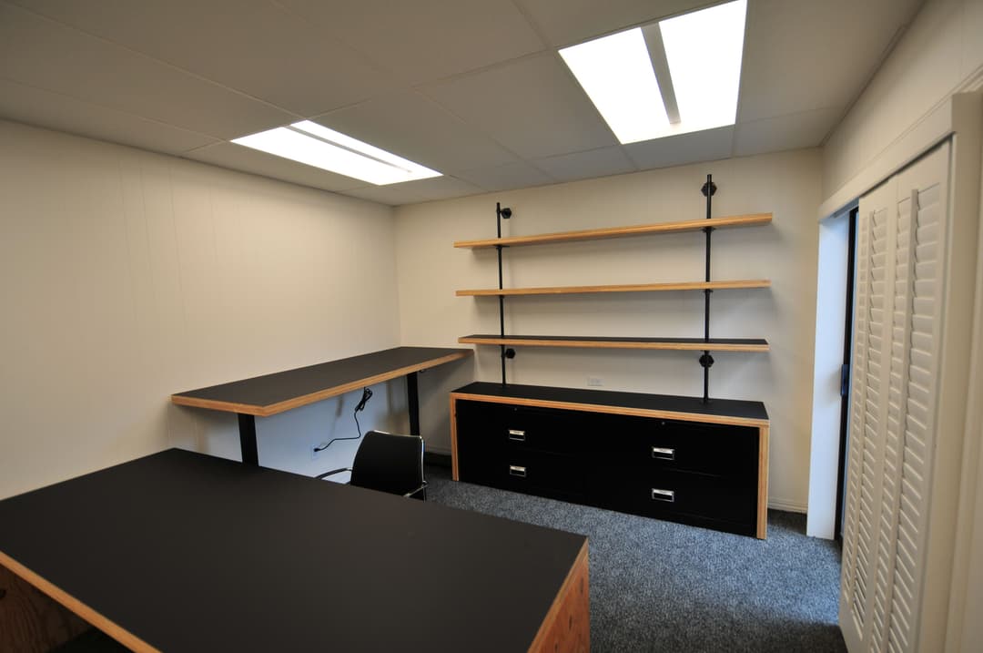 Modern office space featuring black desks, open shelving, and gray carpet flooring.