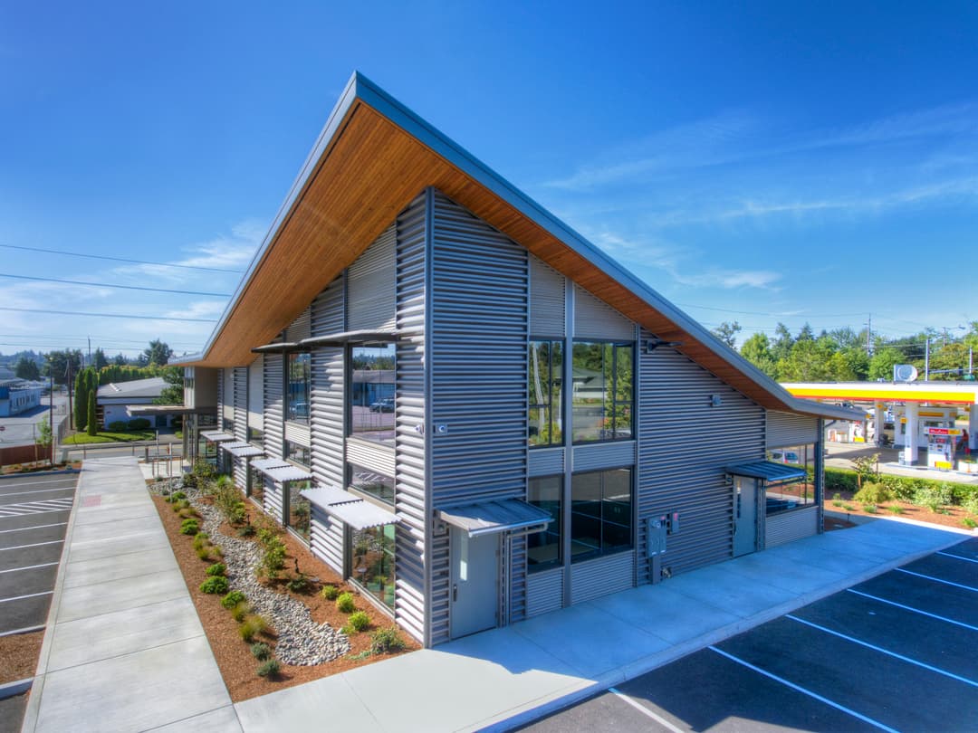 Modern commercial building with large windows and sloped roof, surrounded by landscaped parking area.