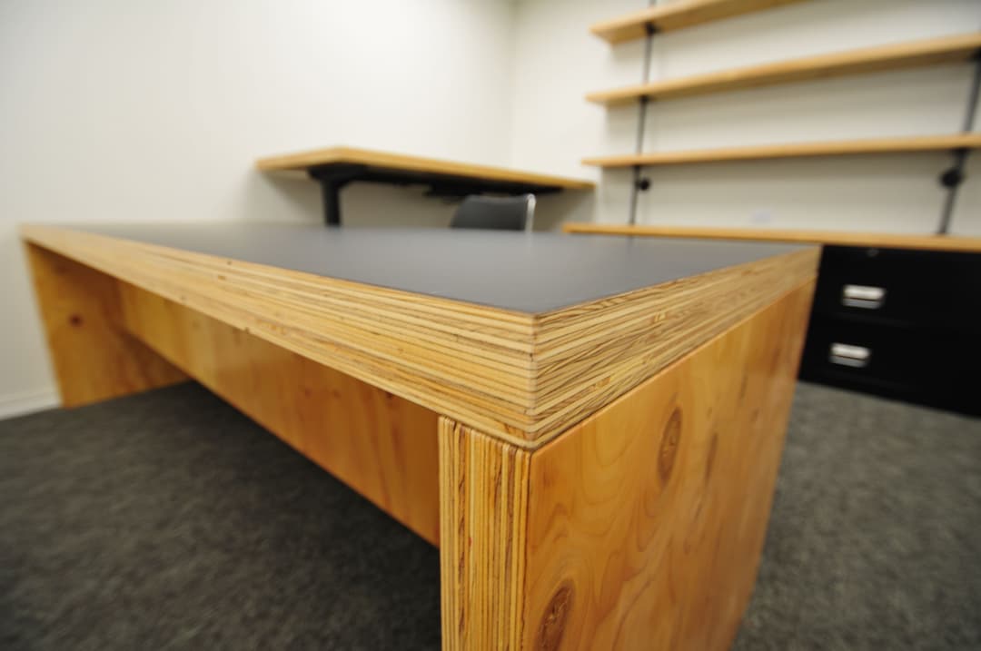Modern wooden desk with a sleek black surface, surrounded by shelving in a minimalist office.