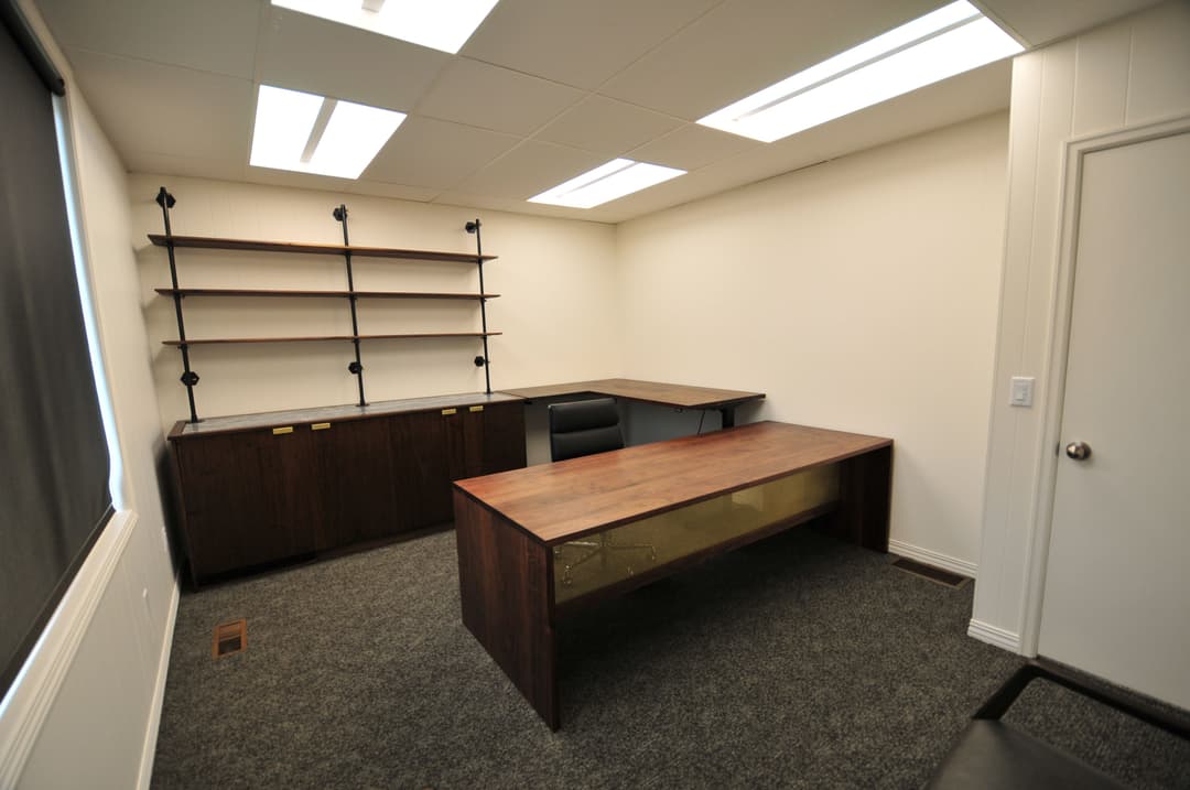 Modern office interior featuring two wooden desks, open shelving, and carpeted flooring.