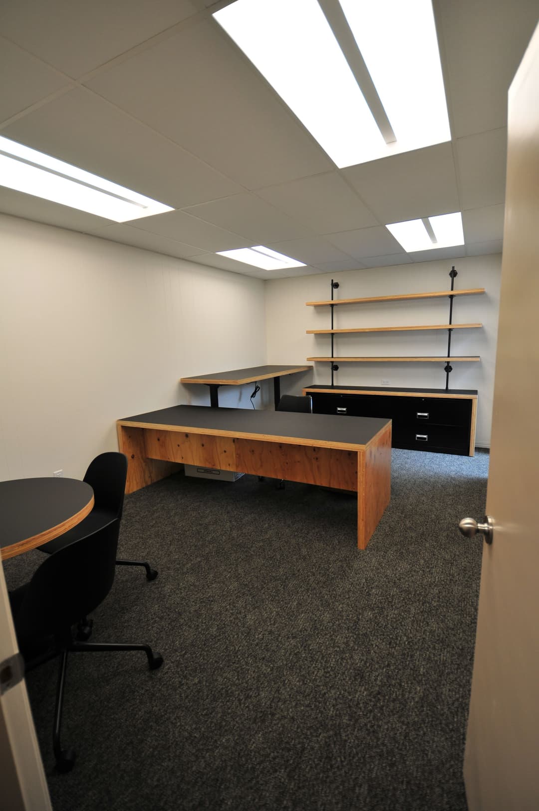 Modern office space with wooden desks, shelves, and carpeted floor, featuring bright lighting.