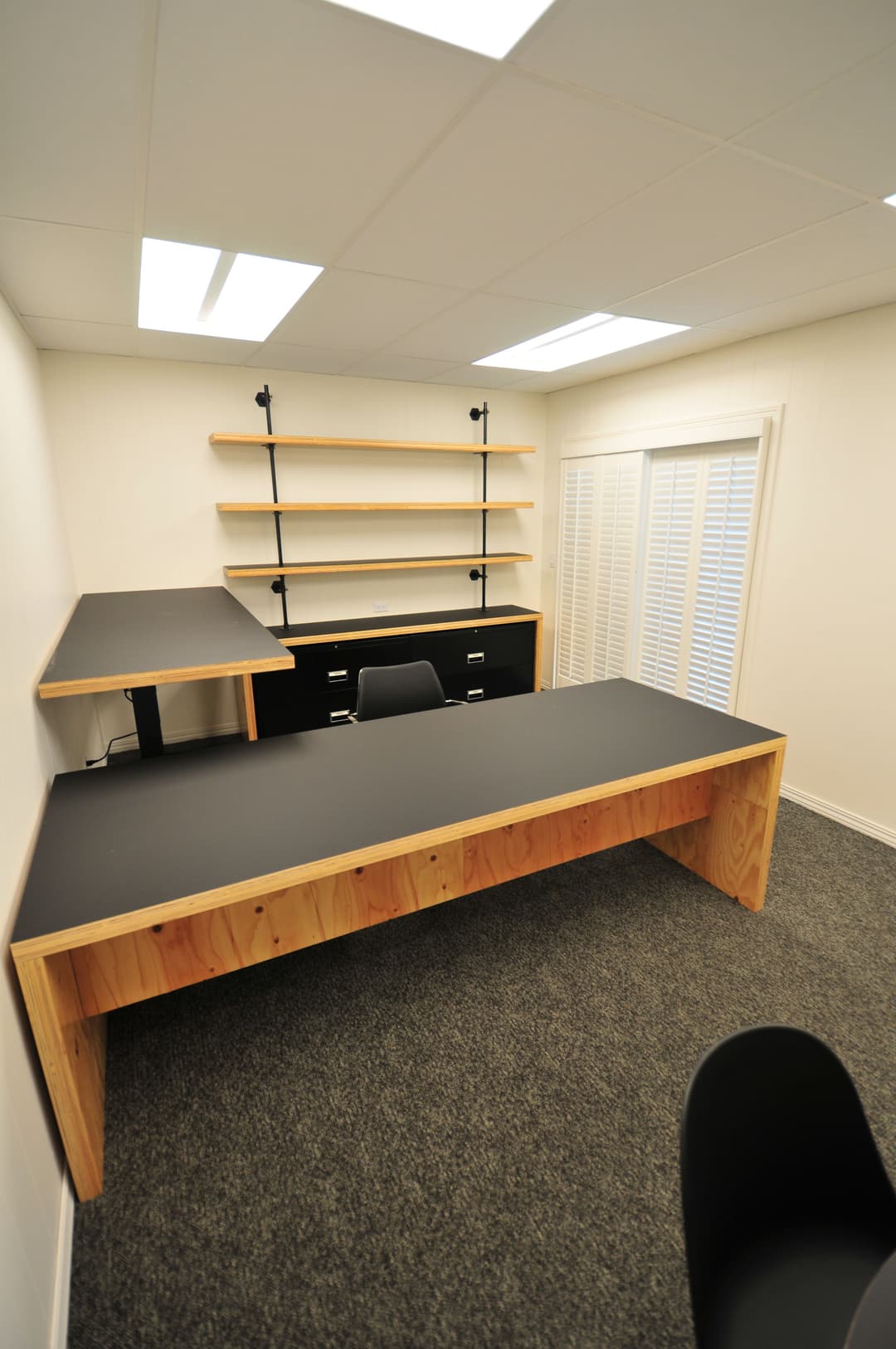 Modern minimalist office space with wooden desk, black shelving, and light gray carpet.
