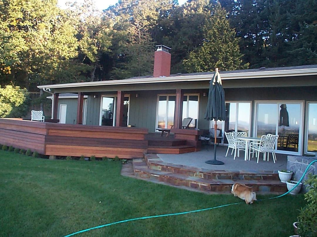 Modern home exterior with wooden deck, patio seating, and grassy yard. Dog in foreground.