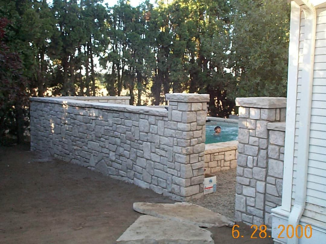 Stone wall enclosing a backyard pool, surrounded by greenery, with a person swimming.