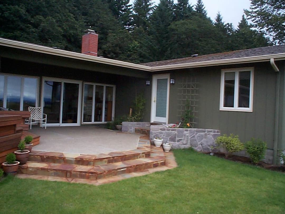 Modern single-story home with stone steps, patio, and green landscape surrounded by trees.