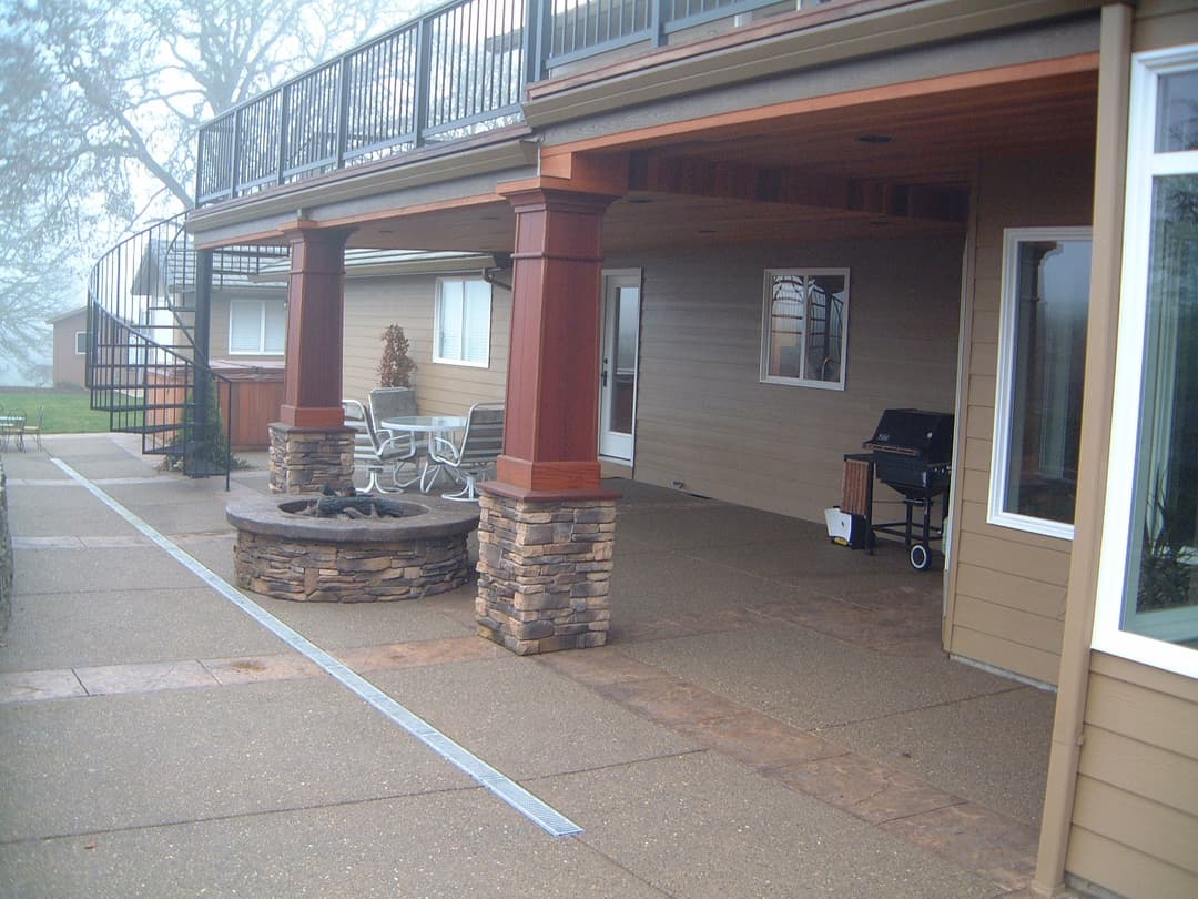 Covered outdoor patio with stone fire pit, grill, and seating area in a misty setting.