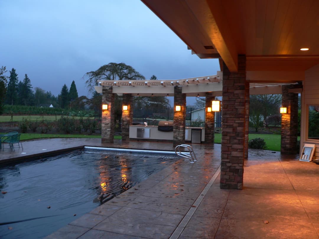 Modern backyard with a pool, outdoor kitchen, stone pillars, and a scenic view.