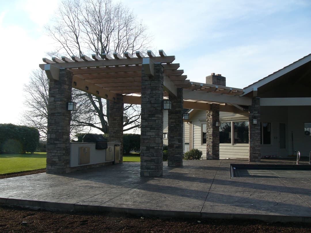 Modern outdoor patio with stone columns and wooden pergola, featuring a grill and landscaped yard.