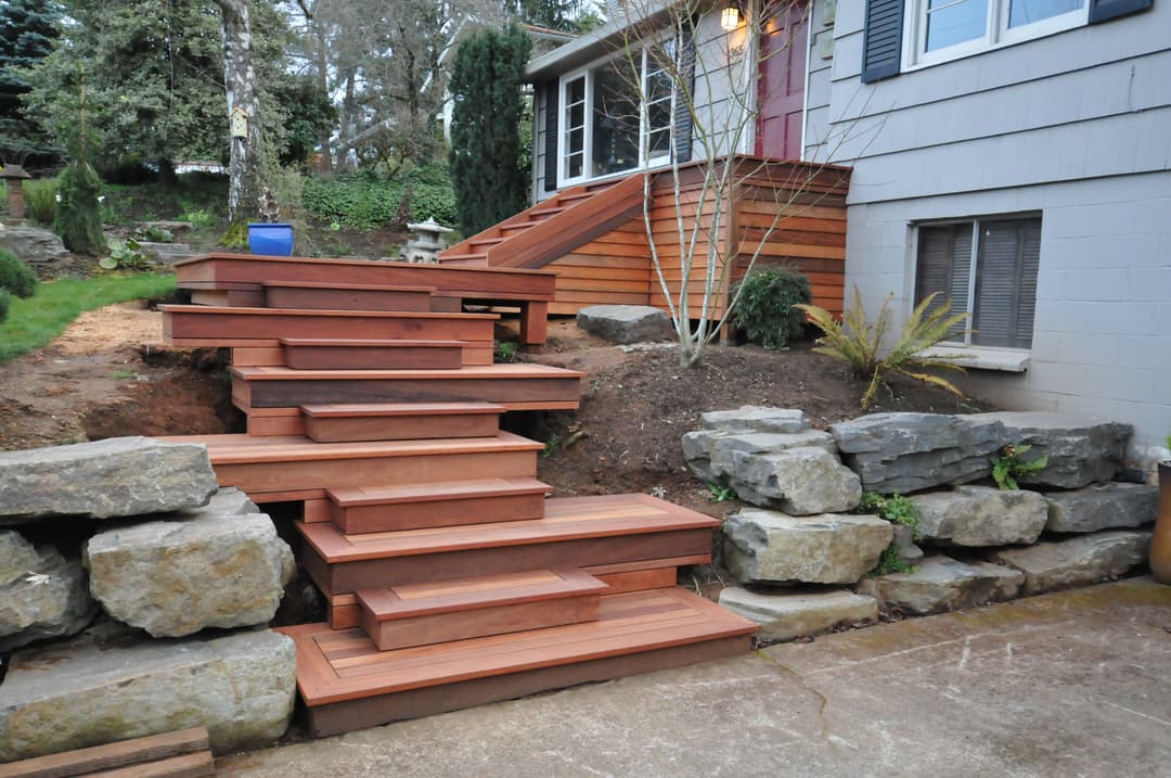 Wooden outdoor steps leading to a house, surrounded by rocks and greenery.