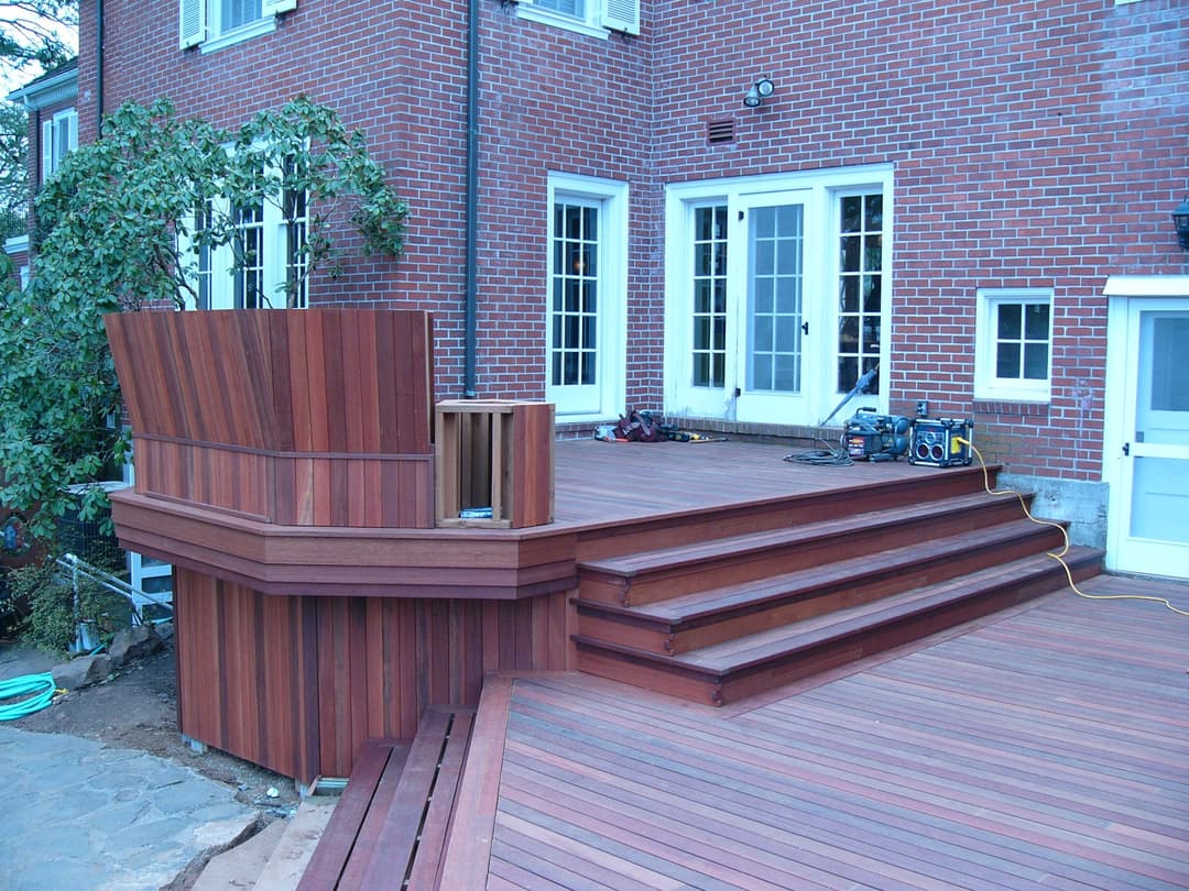 Modern wooden deck with multiple steps and a curved seating area beside a brick house.