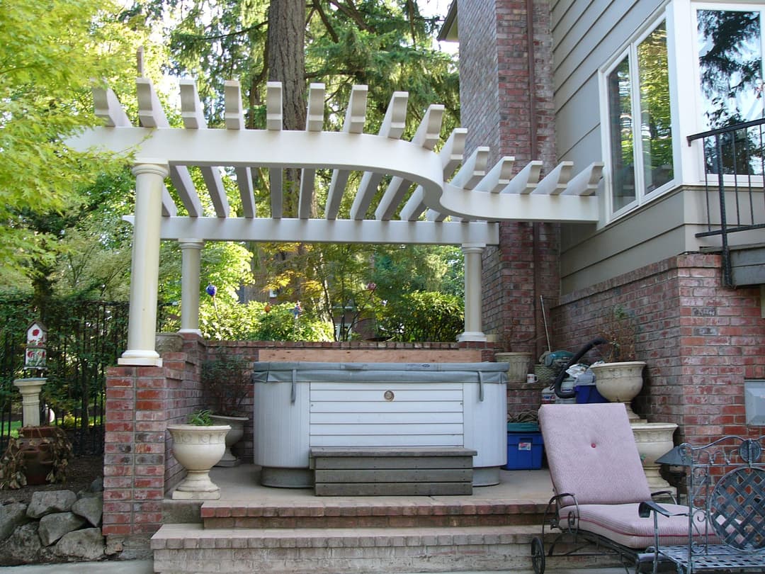 White pergola over a spa area surrounded by greenery and brick walls in a backyard setting.