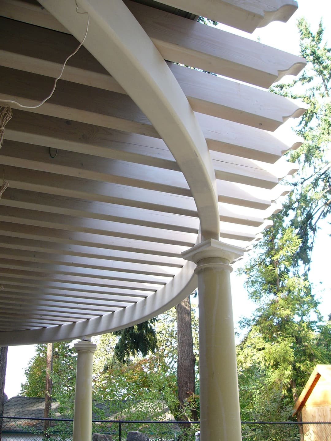 Curved wooden pergola with columns and slatted roof, surrounded by trees.