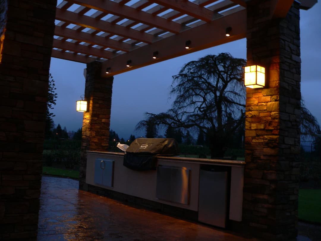 Outdoor kitchen under pergola with stone pillars, grill covered, illuminated by lanterns at dusk.
