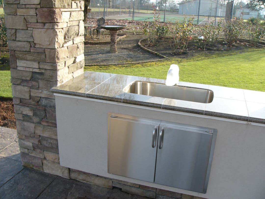 Outdoor kitchen with stone pillars, stainless steel sink, and countertop overlooking landscaped yard.