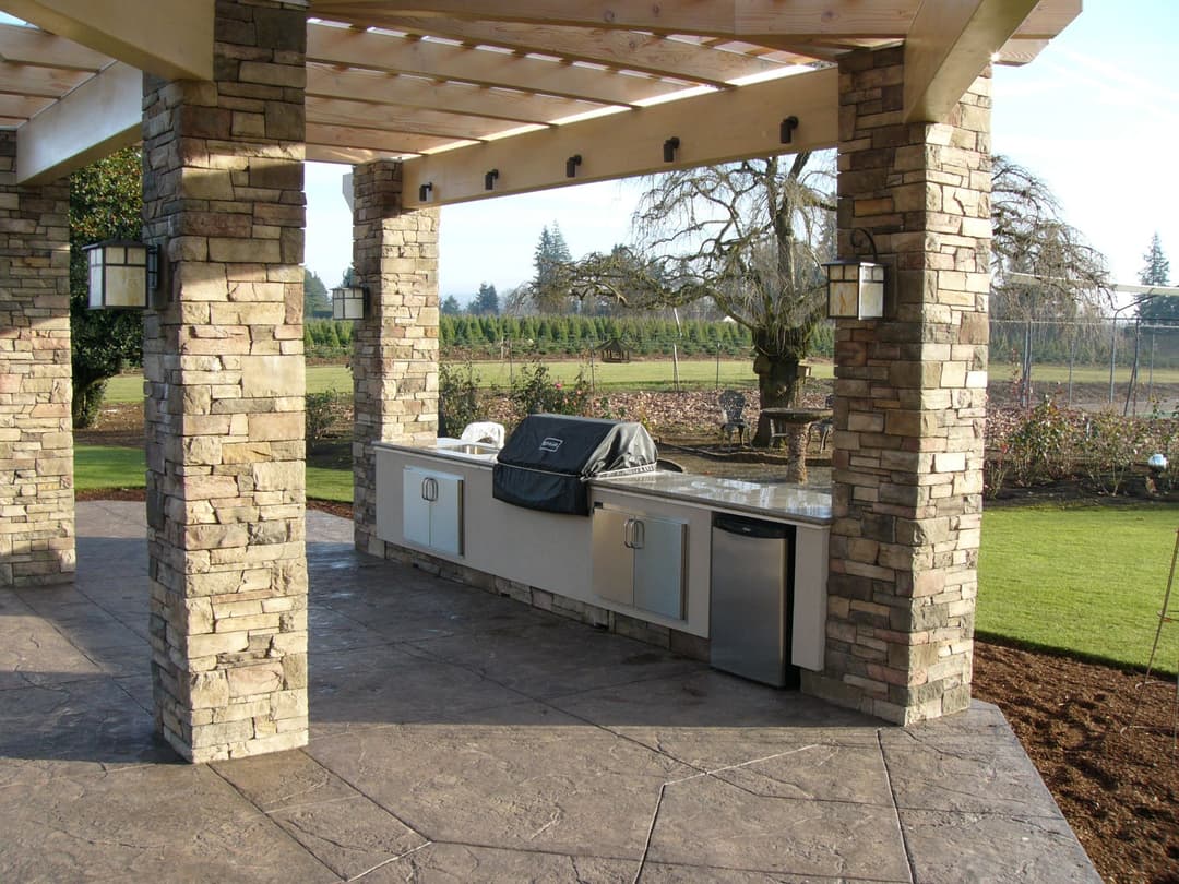 Outdoor kitchen with stone pillars, grill, and countertop in a garden setting.