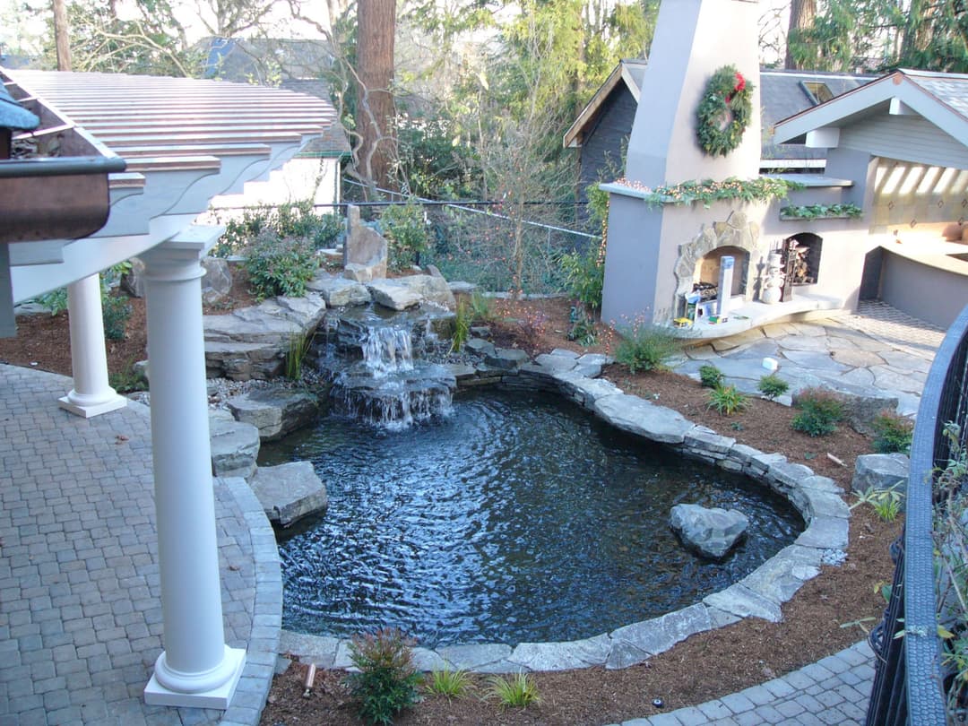 Serene backyard pond with waterfall, stone features, and outdoor kitchen area.