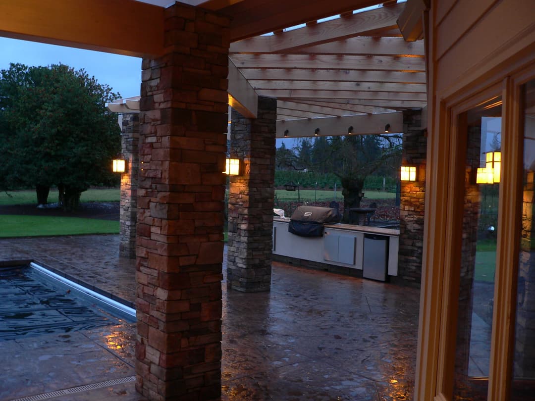 Outdoor patio with stone pillars, kitchen area, pool, and evening lighting in a serene setting.