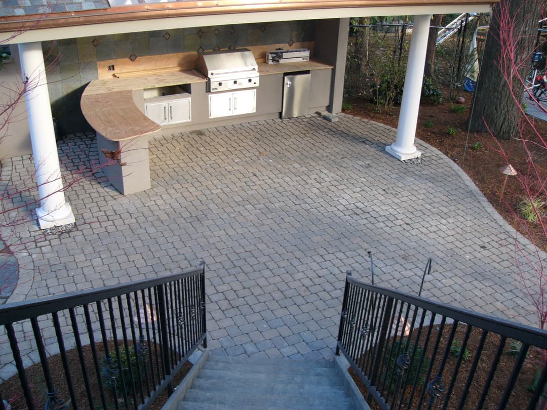 Outdoor kitchen with grill and stone patio, featuring steps and landscaped surroundings.