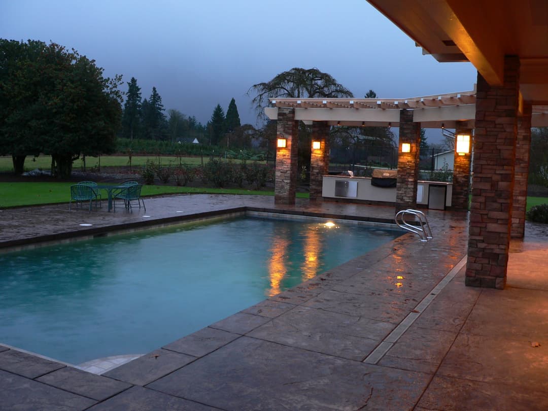 Modern backyard pool with stone patio, cozy lighting, and outdoor kitchen at dusk.