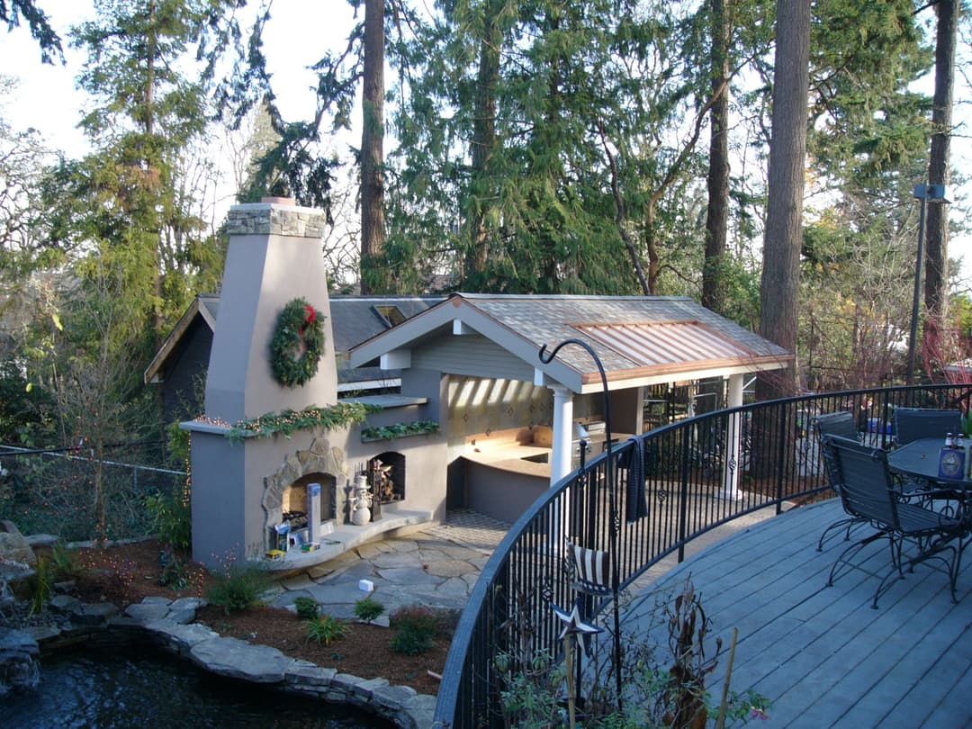 Outdoor kitchen and seating area surrounded by trees, featuring a stone fireplace and festive decor.