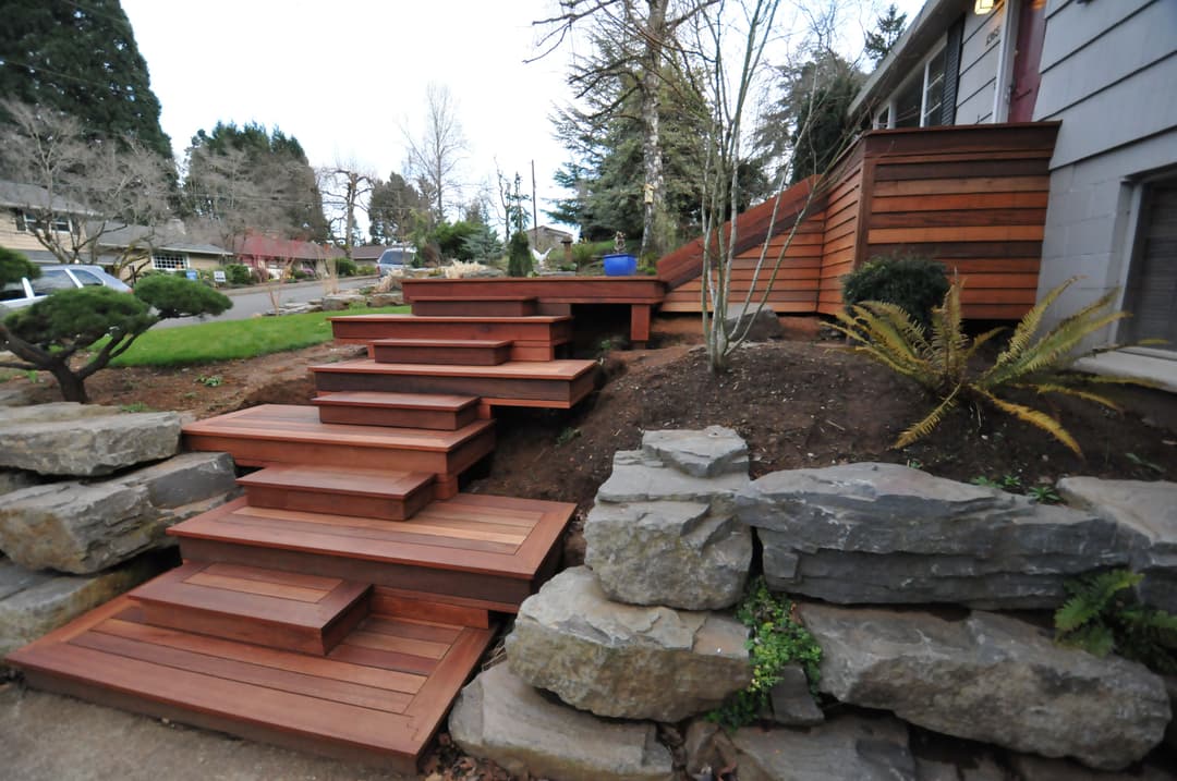 Wooden outdoor steps leading to a house, surrounded by landscaping and stone features.