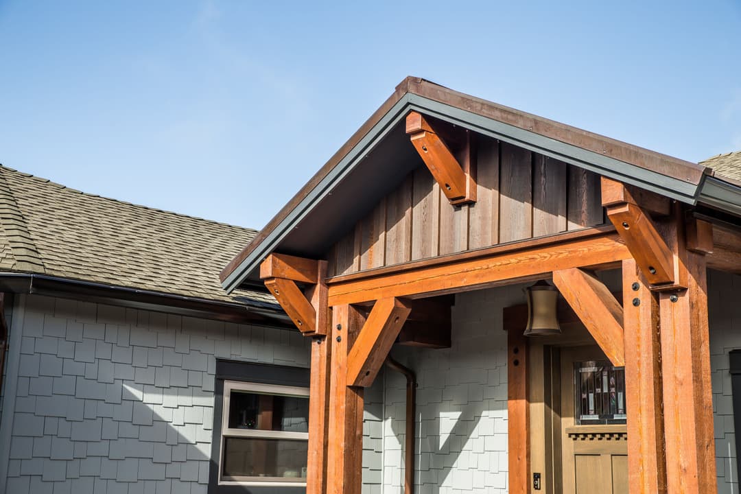 Craftsman-style home entrance with wood beams, shingle siding, and clear blue sky backdrop.