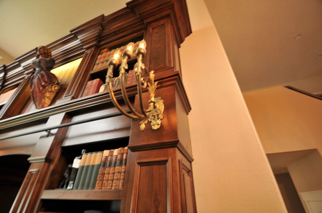 Elegant wooden bookshelf with ornate candle sconces and classic books in a well-lit room.
