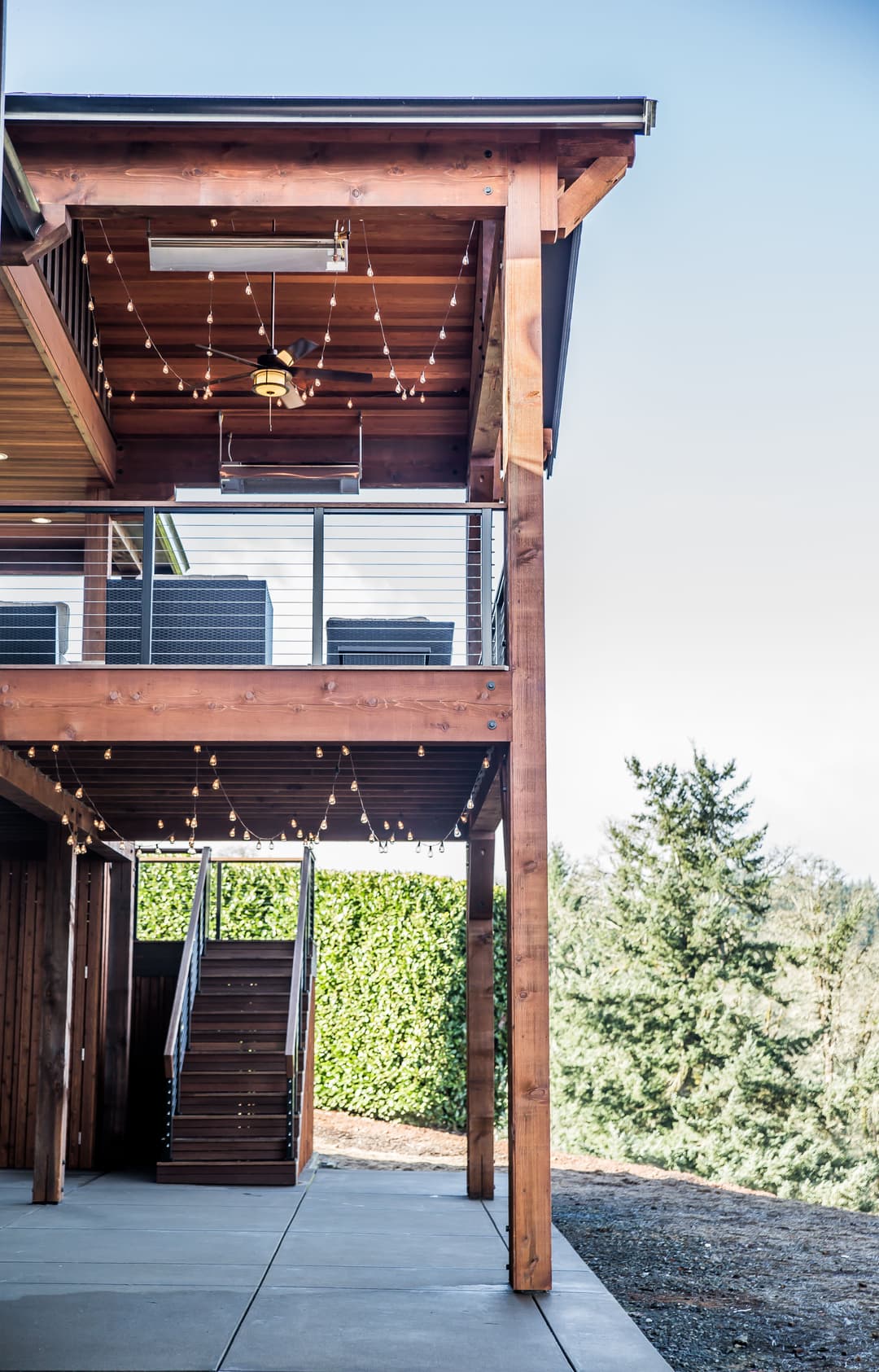 Modern wooden deck with outdoor ceiling fan, string lights, and lush greenery view.