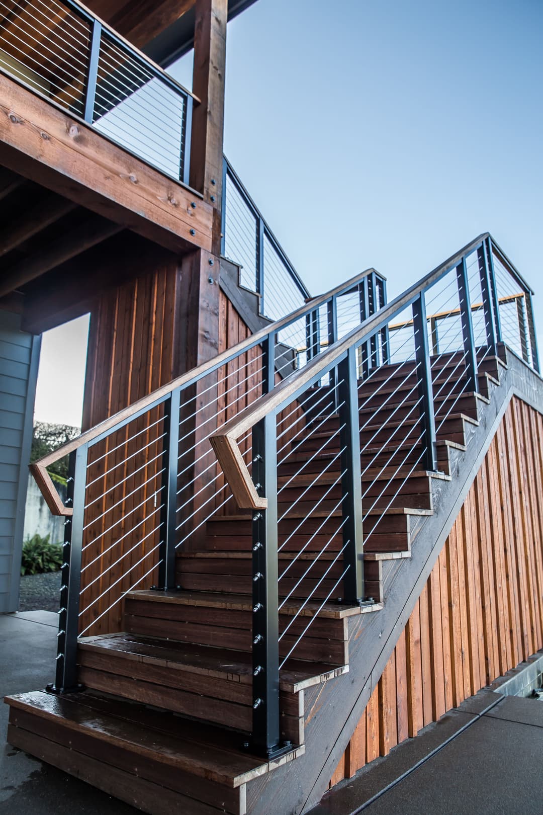 Modern wooden outdoor staircase with black railings and cable mesh detail against a clear sky.