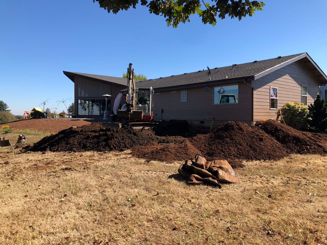 Excavator digging in the yard of a modern house with dirt mounds under a clear blue sky.