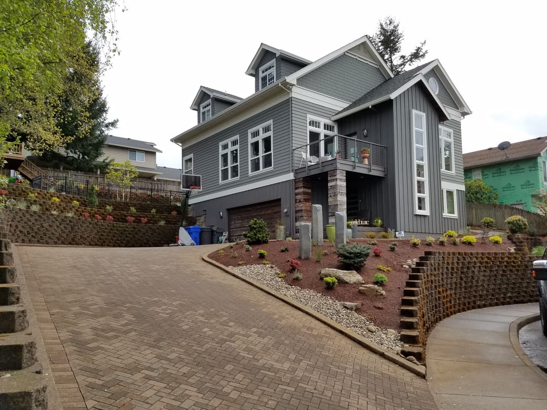 Modern gray house with landscaped yard and driveway on a hill. Basketball hoop visible.