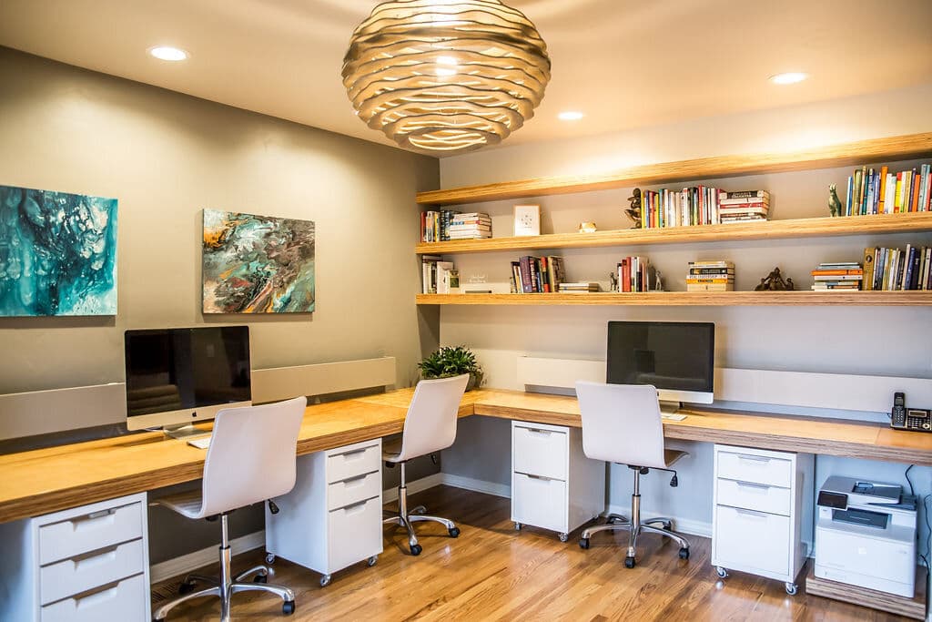 Modern home office with two desks, computers, a bookshelf, and artistic decor. Bright, inviting design.
