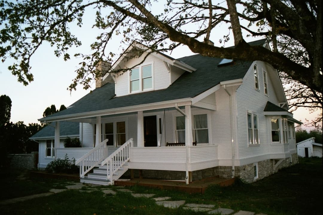 Vintage white house with porch and large tree in front during golden hour.