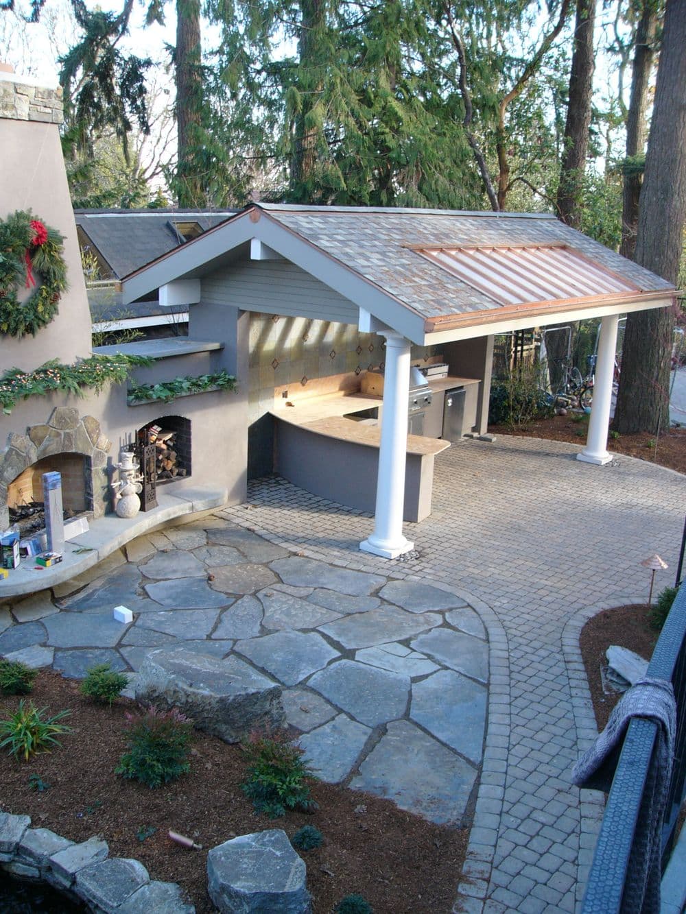 Outdoor kitchen with stone patio, covered area, and grill surrounded by lush greenery.