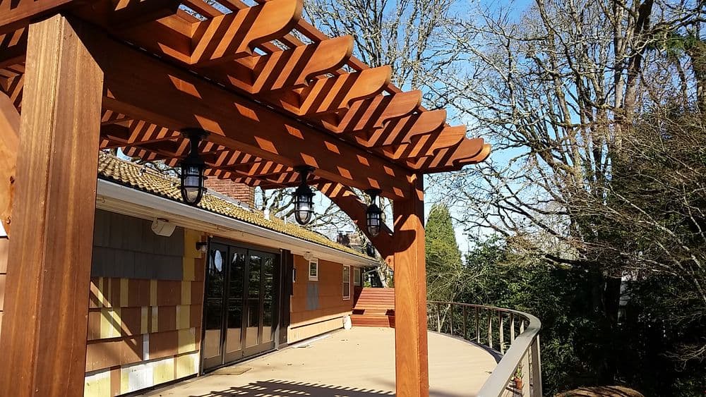 Wooden pergola with lanterns overlooking a sunlit deck and trees.