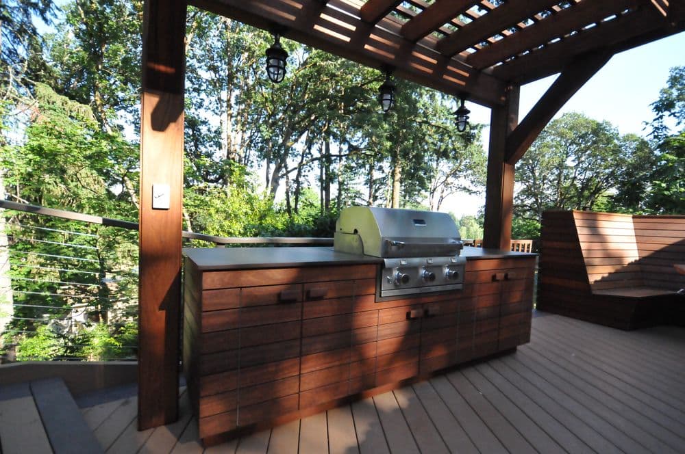 Modern outdoor kitchen with stainless steel grill on a wooden deck surrounded by greenery.