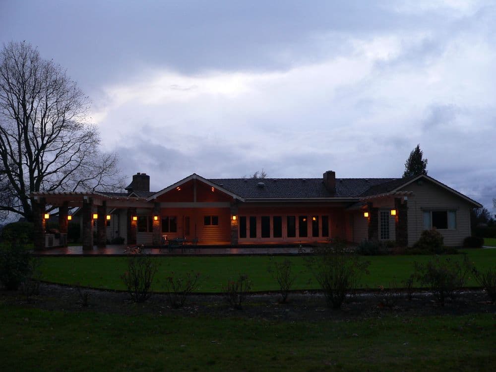 Modern ranch-style house with warm lights, expansive lawn, and overcast sky.