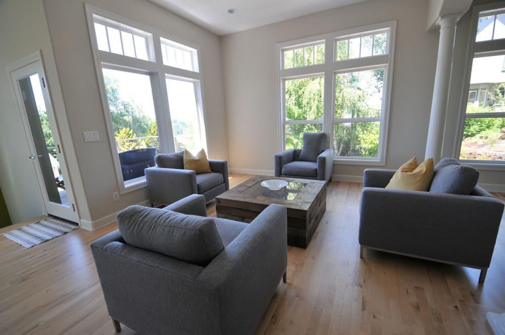 Modern living room with large windows, gray armchairs, a wooden coffee table, and natural light.