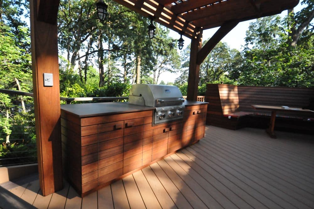 Outdoor kitchen with a grill and wooden seating surrounded by lush greenery.