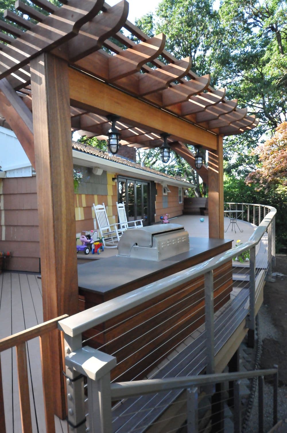 Modern wooden deck with a pergola, grill, and rocking chairs surrounded by greenery.