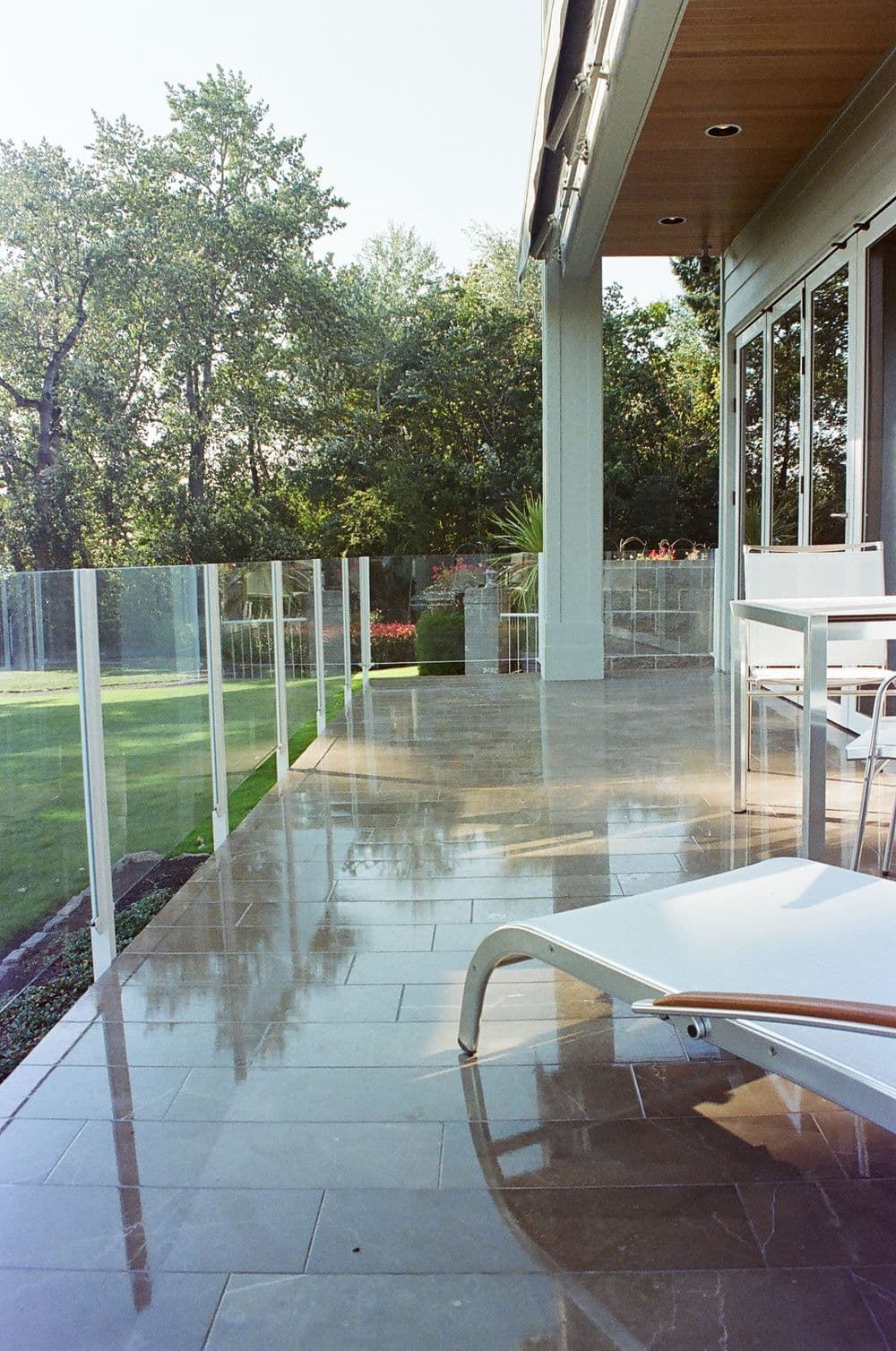 Modern patio with glass railing, sleek tiles, and lounge chair overlooking a lush garden.