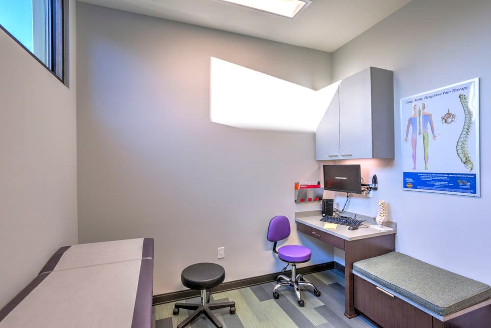 Clean medical exam room with an examination table, desk, computer, and anatomical chart.